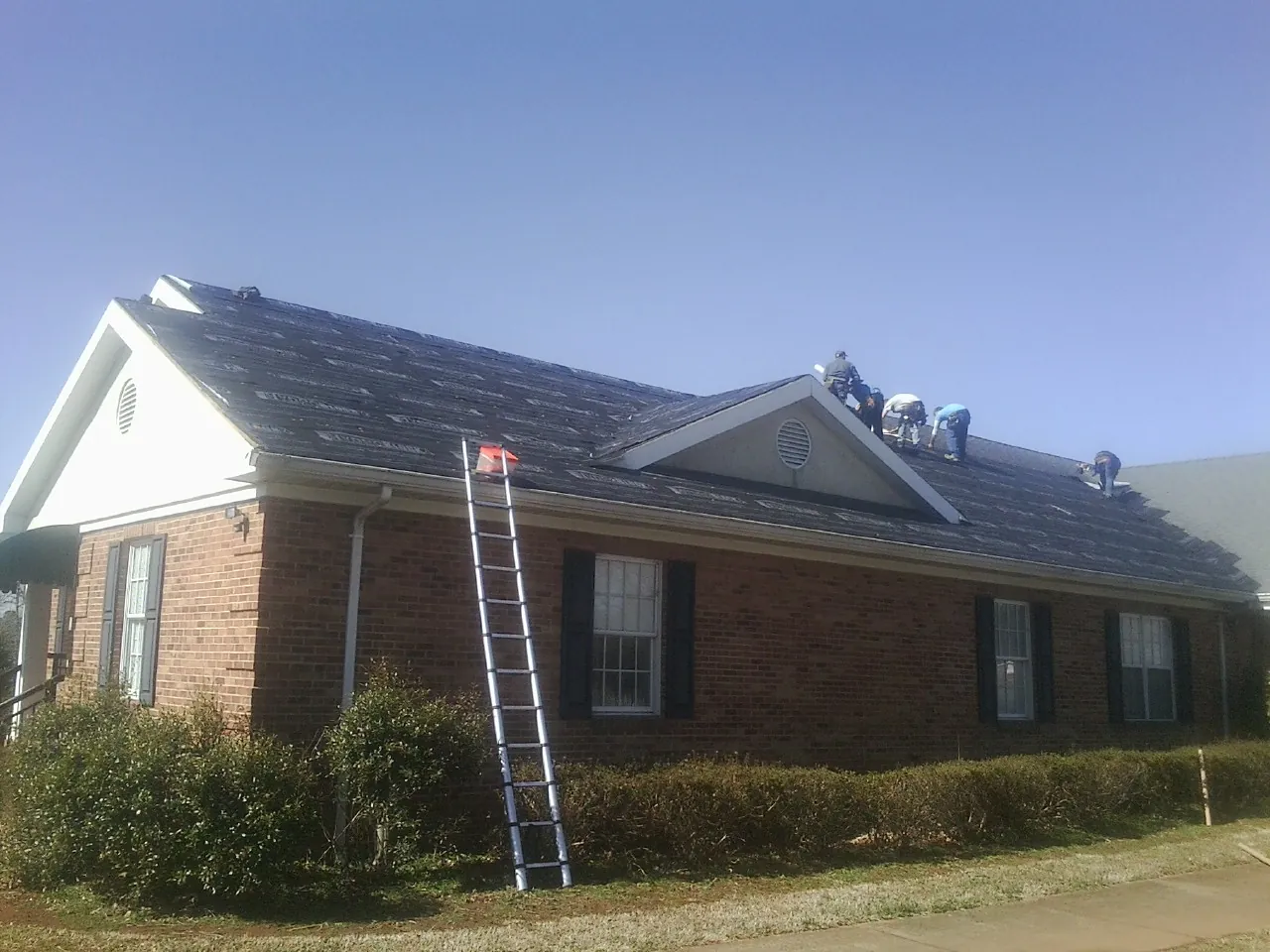 Crew tearing shingles off a brick ranch roof with a ladder set — Metal Roof Repair work by our crew in Fort Ripley